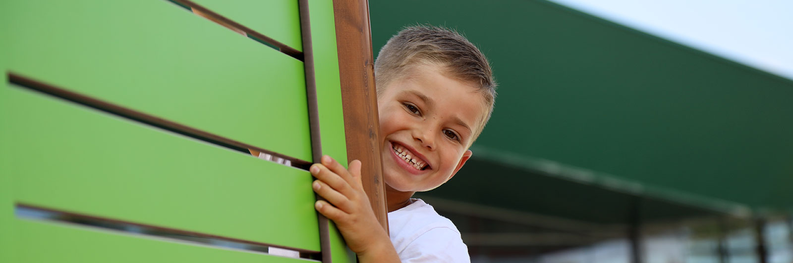 A boy looks around the corner of a playground unit, he is looking at the camera and smiling widely.